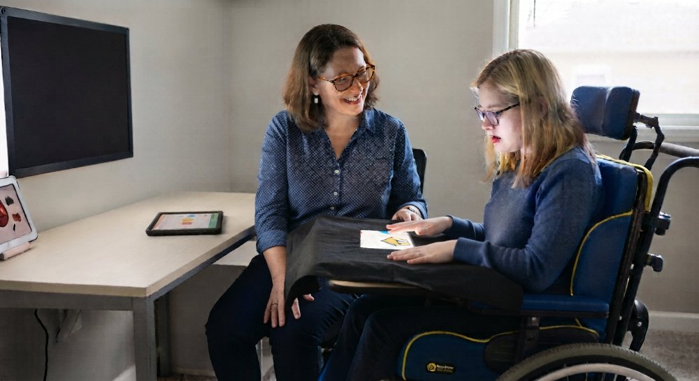 A developmental educator sitting, facing a client in a wheelchair. An AAC device is on the wheelchair tray, which is adapted with a black cloth and a square cut-out to minimise visual distractions.
